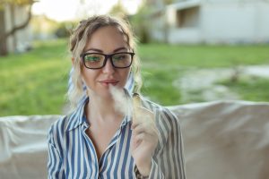 Woman wearing black glasses outdoors, exhaling vapor from a vape pen.