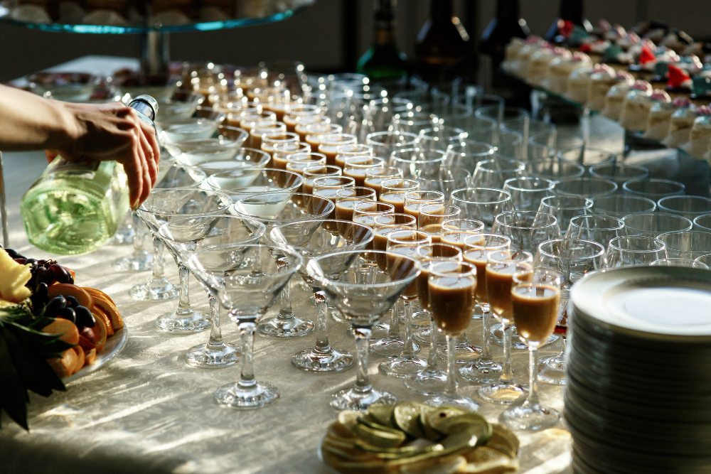 Server pours champagne into dozens of glasses arranged on a banquet table, with fruit and desserts nearby.